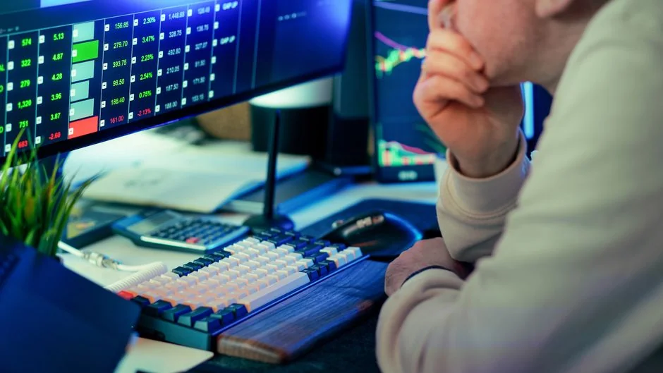 Two business professionals examine stock market figures on a smartphone and laptop in a contemporary office environment.